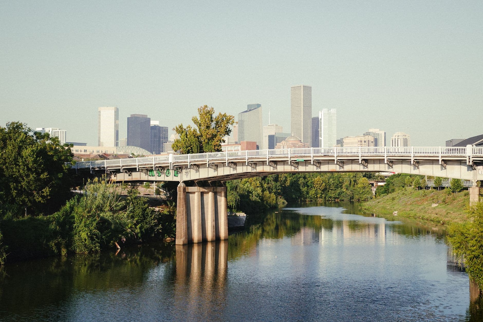 A tranquil view of Houston's skyline with a bridge over a serene bayou, surrounded by lush greenery.