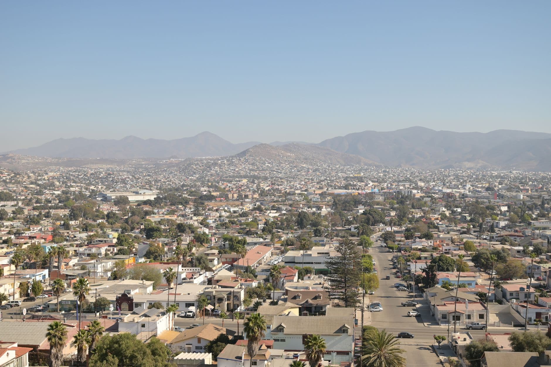 Aerial daytime view of Tijuana's cityscape with mountainous backdrop under a clear sky.